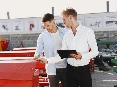 Two men discussing agricultural equipment purchase at a dealership, one holding a clipboard outside.